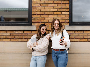 Two women standing against a brick wall smile while looking at a phone together; one holds a laptop and the other carries a backpack.