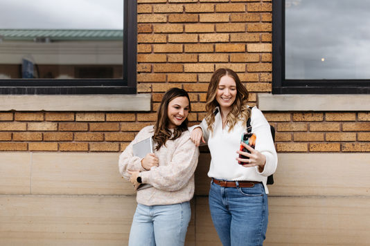 Two women standing against a brick wall smile while looking at a phone together; one holds a laptop and the other carries a backpack.