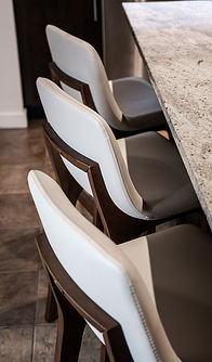 Kitchen counter seating with white upholstered leather stools, stone countertop, warm wood frames, and neutral finishes.