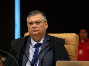 Imagem de um homem sentado em uma cadeira de tribunal, usando traje formal e óculos, com uma expressão séria. Ele está em uma sala de julgamento, com outros elementos de fundo desfocados.