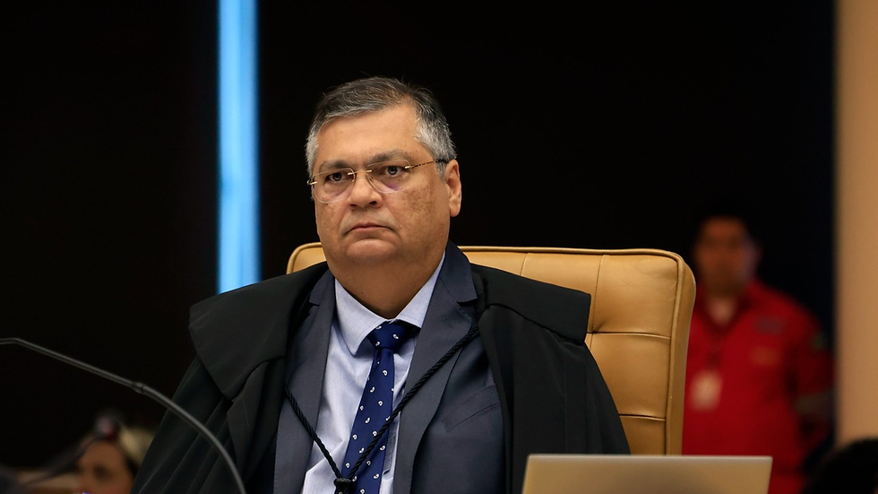 Imagem de um homem sentado em uma cadeira de tribunal, usando traje formal e óculos, com uma expressão séria. Ele está em uma sala de julgamento, com outros elementos de fundo desfocados.
