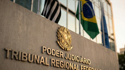 Fachada do Tribunal Regional Federal (Poder Judiciário), com o brasão dourado e bandeiras ao fundo, incluindo as do Brasil e de São Paulo.