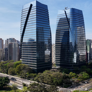 Vista urbana de dois edifícios corporativos espelhados e modernos, lado a lado, cercados por árvores e avenidas, com diversos prédios altos ao fundo sob céu claro.