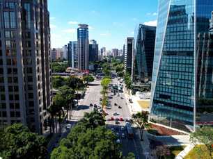 Vista aérea de avenida larga em centro urbano, cercada por edifícios altos e modernos de vidro, com trânsito leve, árvores ao longo da via e céu azul ao fundo.