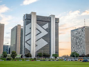 Prédio corporativo em Brasília com fachada espelhada e formas geométricas, em área gramada e cercado por outros edifícios ao entardecer.