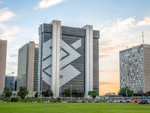 Prédio corporativo em Brasília com fachada espelhada e formas geométricas, em área gramada e cercado por outros edifícios ao entardecer.
