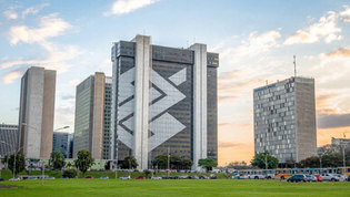 Prédio corporativo em Brasília com fachada espelhada e formas geométricas, em área gramada e cercado por outros edifícios ao entardecer.