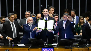 Grupo de autoridades em plenário legislativo. No centro, um homem sorri e ergue um documento aberto, enquanto outros políticos ao redor observam; à esquerda, um homem de barba branca ajuda a segurar o papel, e à direita outro homem faz um gesto com a mão. Ao fundo, várias pessoas acompanham a cena em pé.