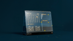 Calendário de mesa de acrílico com design moderno e minimalista, exibindo o mês de maio de 2026. O layout inclui áreas destacadas para anotações como “aniversários” e “não esquecer”, além de mini calendários de meses adjacentes na base. O objeto está posicionado sobre uma superfície escura, com detalhes em dourado que reforçam um visual sofisticado.