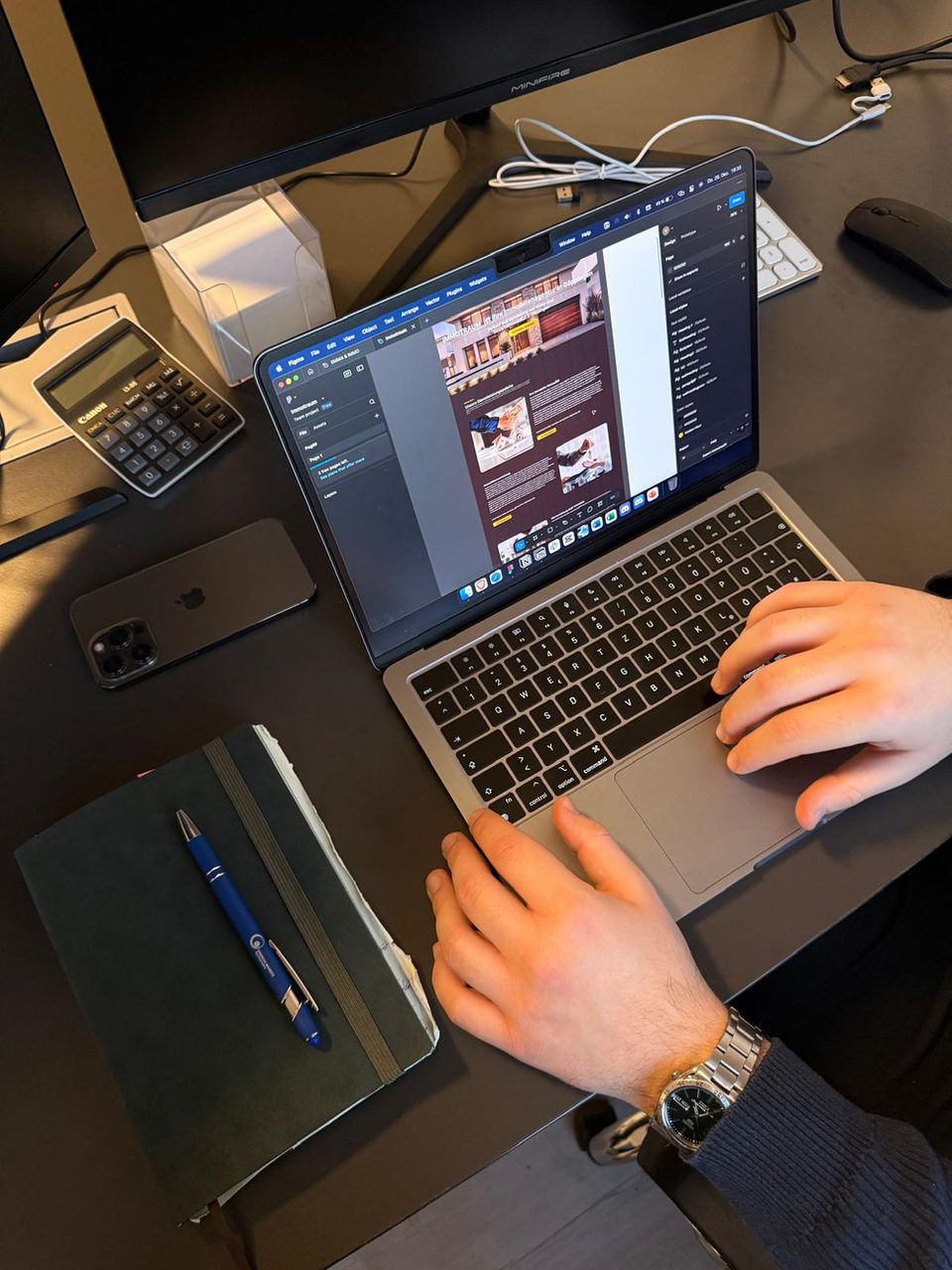 Zwei Monitore im Hintergrund und ein Macbook mit Notizblock daneben, auf dem Macbook ist ein Projekt von Artistic Avenue geöffnet