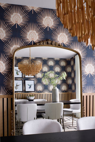 Dining room mirror reflecting bold navy and cream wallpaper, gold chandelier, white dining chairs, and wood paneling.