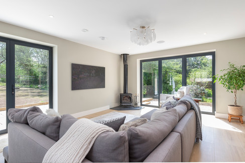 Modern living room featuring a grey sectional sofa, freestanding wood-burning stove, minimalist wall art, and floor-to-ceiling glass doors overlooking a garden.
