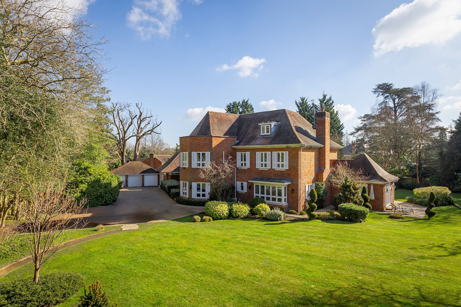 Large detached brick house with landscaped gardens and mature trees, photographed from elevated angle on a sunny day