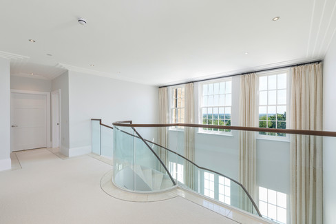 Light-filled hallway landing with full-height sash windows, cream carpets and curved glass staircase overlooking countryside views in upscale British property