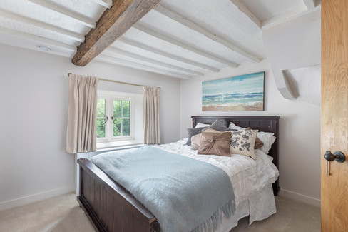 Rustic cottage bedroom with exposed ceiling beams, wooden bed frame, neutral bedding, and small window overlooking greenery