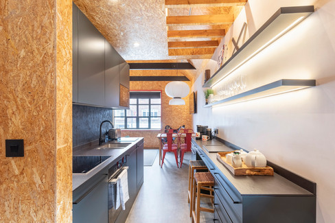 View through galley kitchen towards dining area with red chairs, pendant lighting and plywood finishes