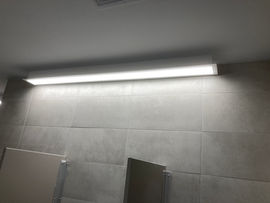 Long fluorescent light fixture illuminates grey tiled wall above bathroom stalls.