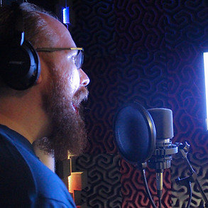 Actor Josh Putnam in profile from the back right, stands in front of a microphone in a recording booth. Josh is smiling, wearing headphones, and smiling. Josh has a beard and glasses, and there is a screen shining light onto him.