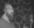 Actor Josh Putnam in profile from the right, stands in front of a microphone in a recording booth. Josh is smiling, wearing headphones, and smiling. Josh has a beard and glasses, and picture is in black and white.