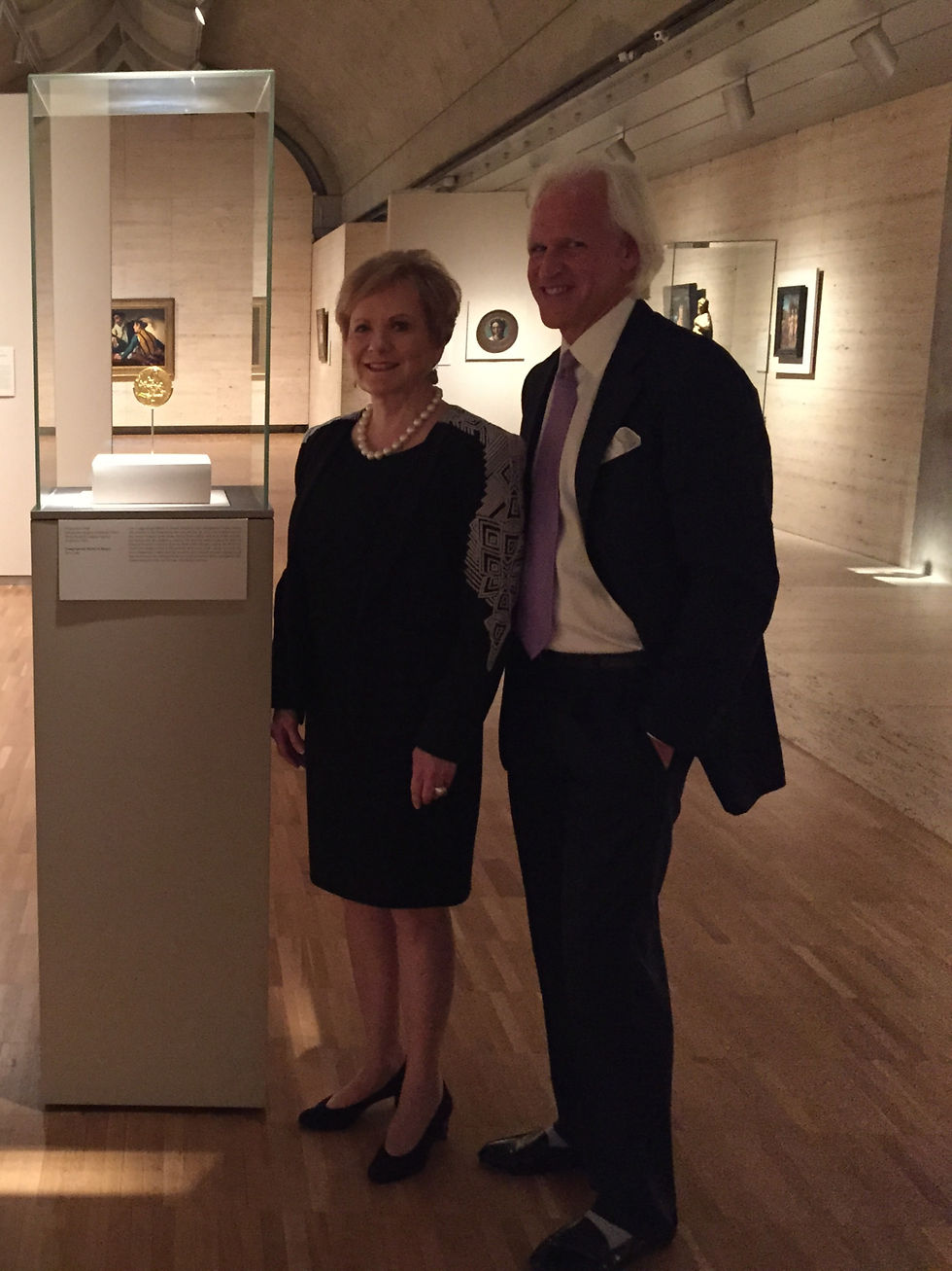Two smiling older adults in formal wear stand beside a glass display case with a gold artifact in a museum gallery.