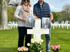 Couple stands at a military grave marker in a cemetery, holding old photos, with white crosses and flower vases; memorial mood.