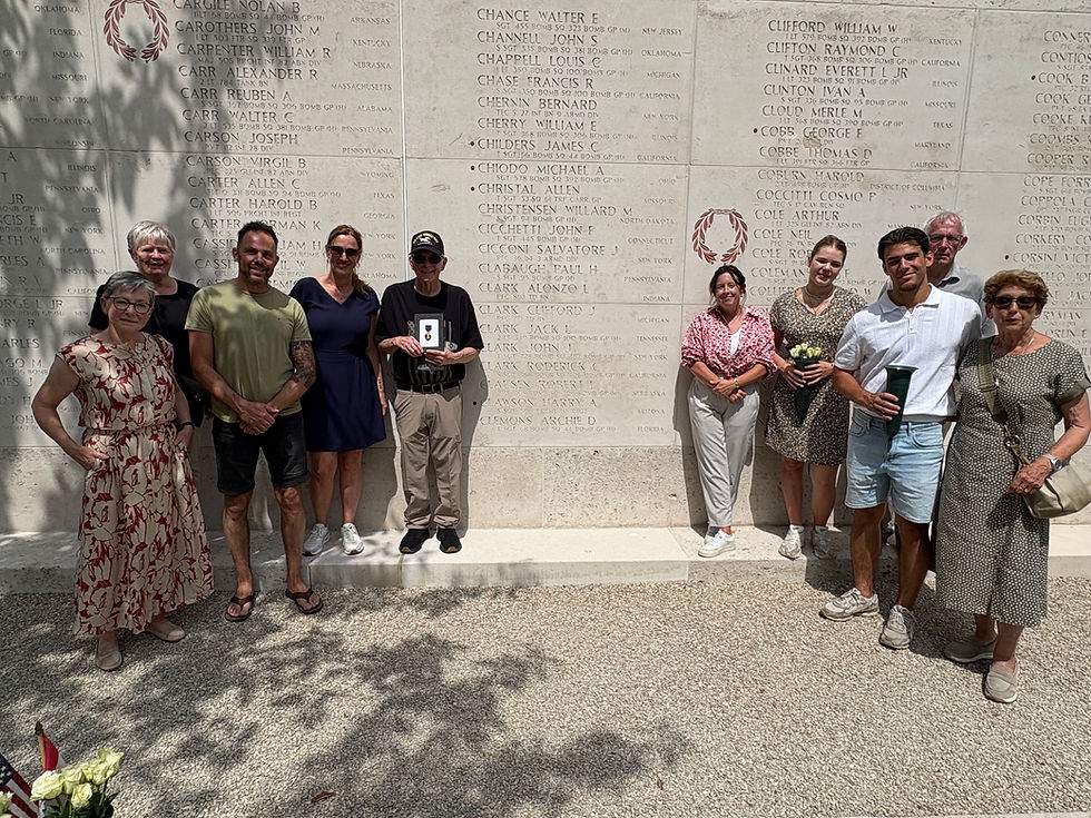 Group poses at an engraved memorial wall; one man holds a medal case while others carry flowers in bright sun.
