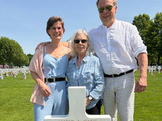 Three smiling adults pose at a cemetery cross grave marked ALFRED A. WANG, with flowers and small flags under a bright blue sky.