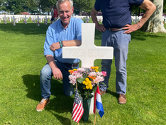 Two men at a military grave marker with flowers and U.S. and Dutch flags in a sunny cemetery, honoring Gordon L. Springsteen.