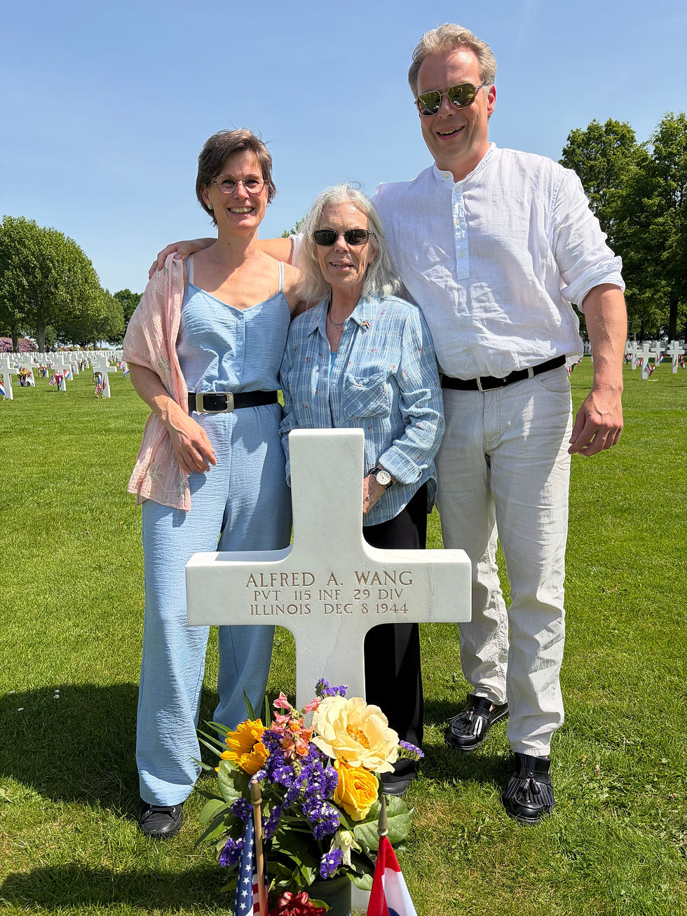 Three smiling adults pose at a cemetery cross grave marked ALFRED A. WANG, with flowers and small flags under a bright blue sky.