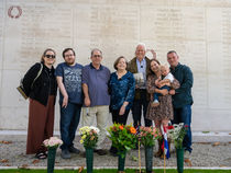 A group of people pose in front of a memorial wall with flowers placed before it. They are smiling. The background features names engraved in stone.
