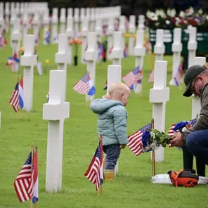 Man kneels to place flowers by white crosses with flags in a cemetery. A child stands nearby. Somber mood on a grassy field.