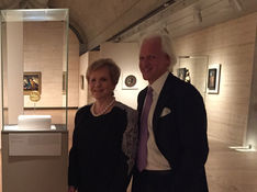 Two smiling older adults in formal wear stand beside a glass display case with a gold artifact in a museum gallery.