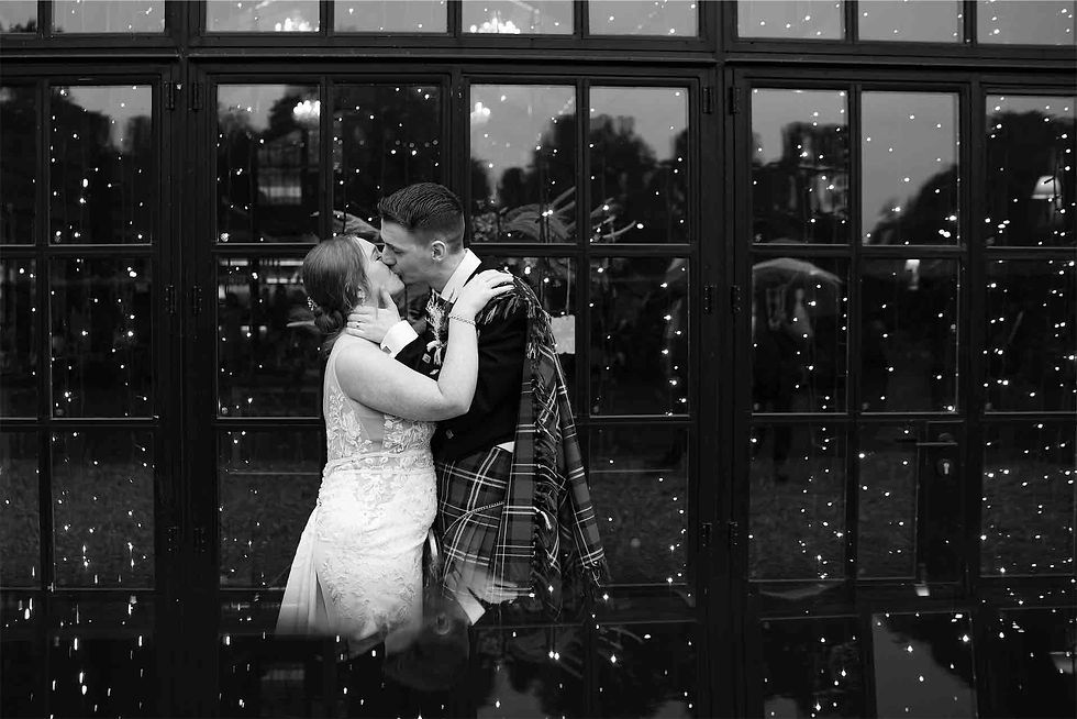 Bride and groom share a kiss in front of a window with string lights. Groom in tartan, bride in lace. Romantic, intimate setting.