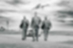 Three men in suits walk confidently on a path by the beach, with a vintage car behind them under a dramatic cloudy sky.
