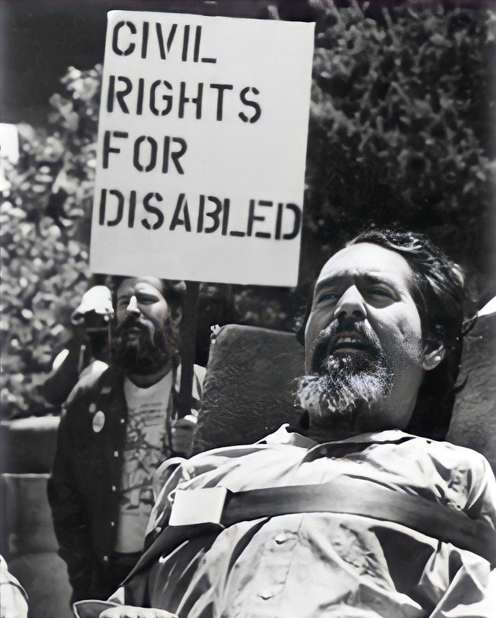 a white man in a wheelchair with a goatee. Behind him a man holds a protest plaque saying "civil rights for disabled"