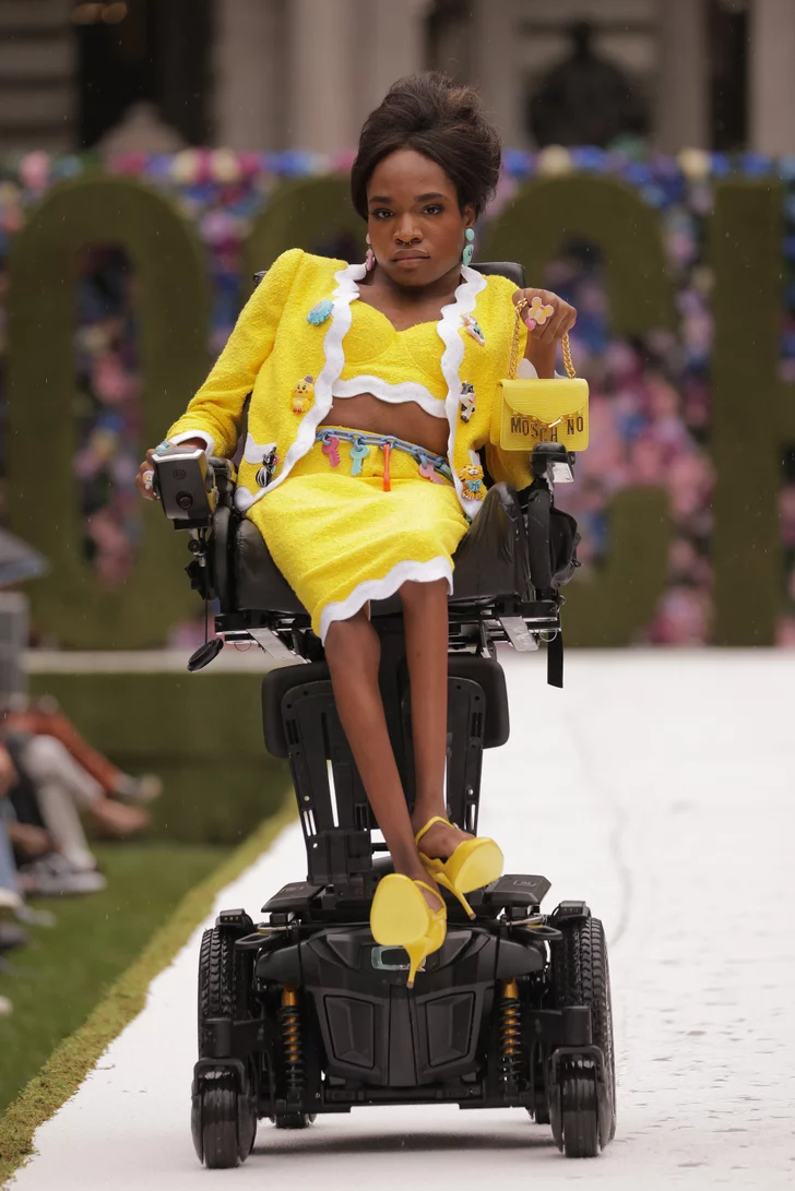 a black transgender woman in a wheelchair with a beehive hairstyle works the runway in a yellow matching skirt suit set and handbag