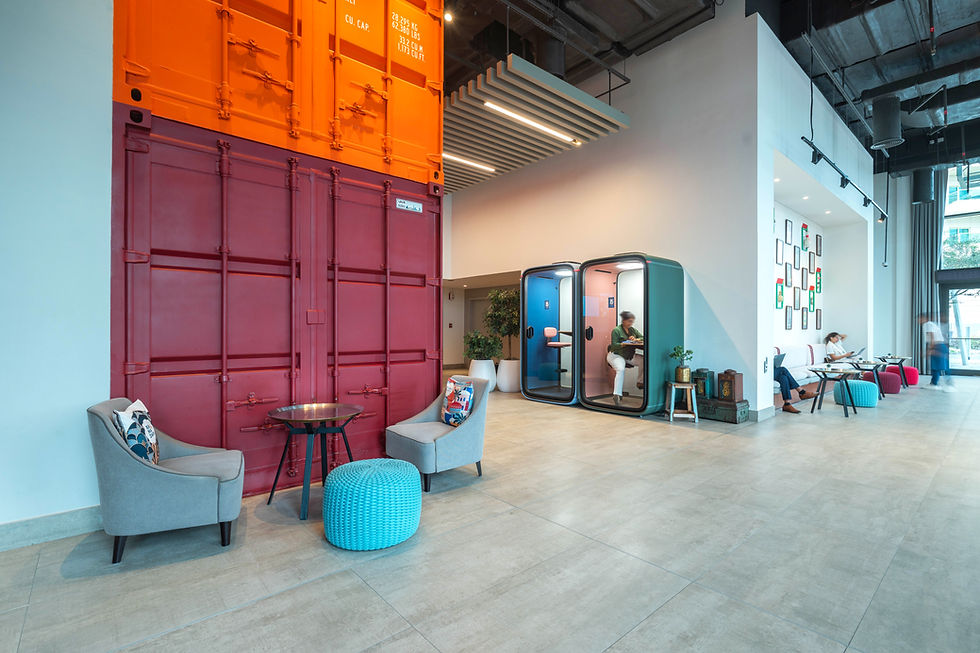 Modern space with shipping container decor; people work in glass pods and lounge in seating area. Bright colors and creative ambiance.
