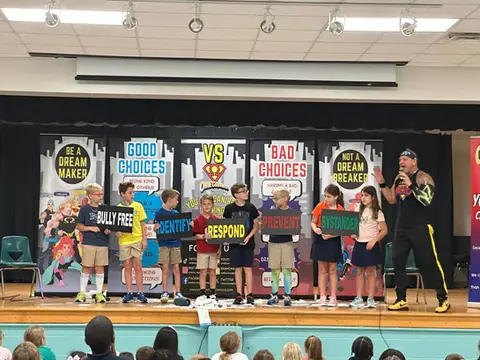 Omegaman leading a school assembly with kids on stage holding signs about making good choices.