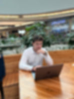 A young man in a white shirt sits at a wooden table, concentrating and working on a laptop. He looks thoughtful and is surrounded by a relaxed environment with plants in the background.