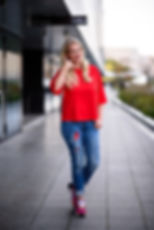 Jessica Lackner in rotem Oberteil und Jeans, freundlich lächelnd mit Telefon-Geste auf einem modernen Gehweg.