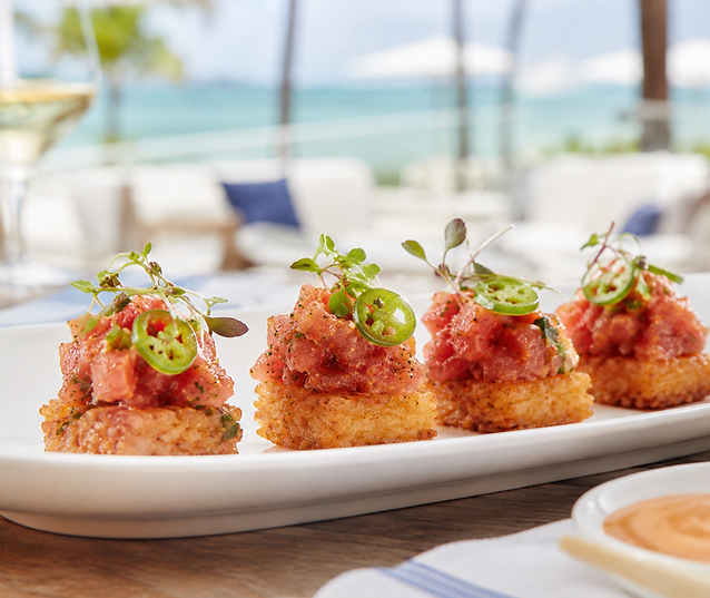 A plate of tuna tartare on toasted bread with microgreens and jalapeño, near a beachside setting.