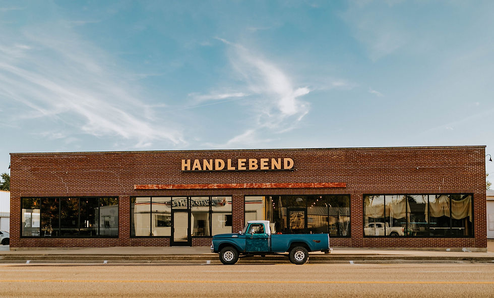 Red brick building with "HANDLEBEND" sign, under a blue sky. A vintage blue truck is parked in front. Large windows reflect street view.