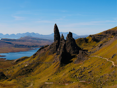 Jagged dark rock spires rise over green hills and a blue loch under a clear sky, with tiny hikers on a winding trail.