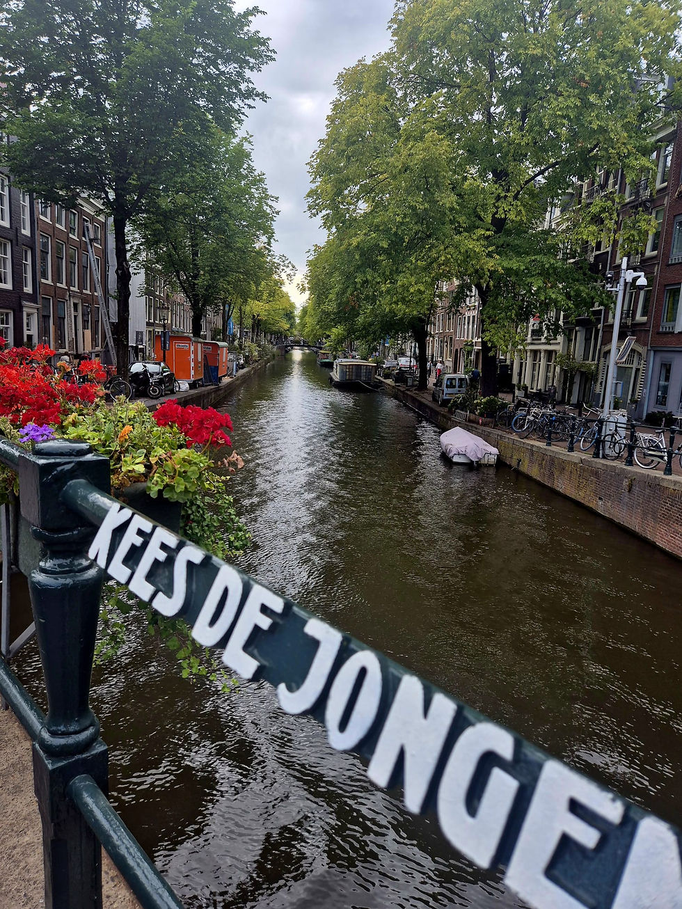 Tree-lined canal between brick houses and bikes, with flowers on a bridge rail and blurred KEES DE JONGE tape in foreground.