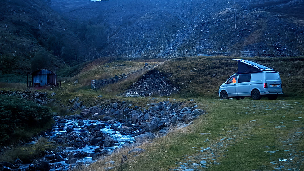 White camper van parked by a rocky stream in a blue-toned mountain valley at dusk, with a quiet, remote mood.
