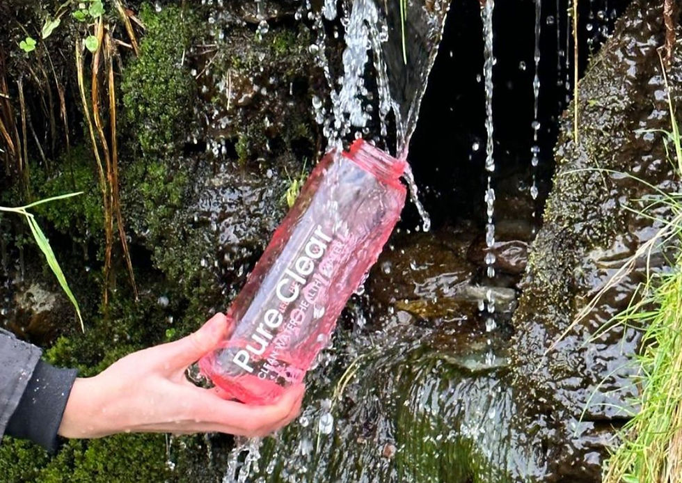 Hand holds a Pure Clear water bottle under a trickling mossy waterfall in a rocky green stream.