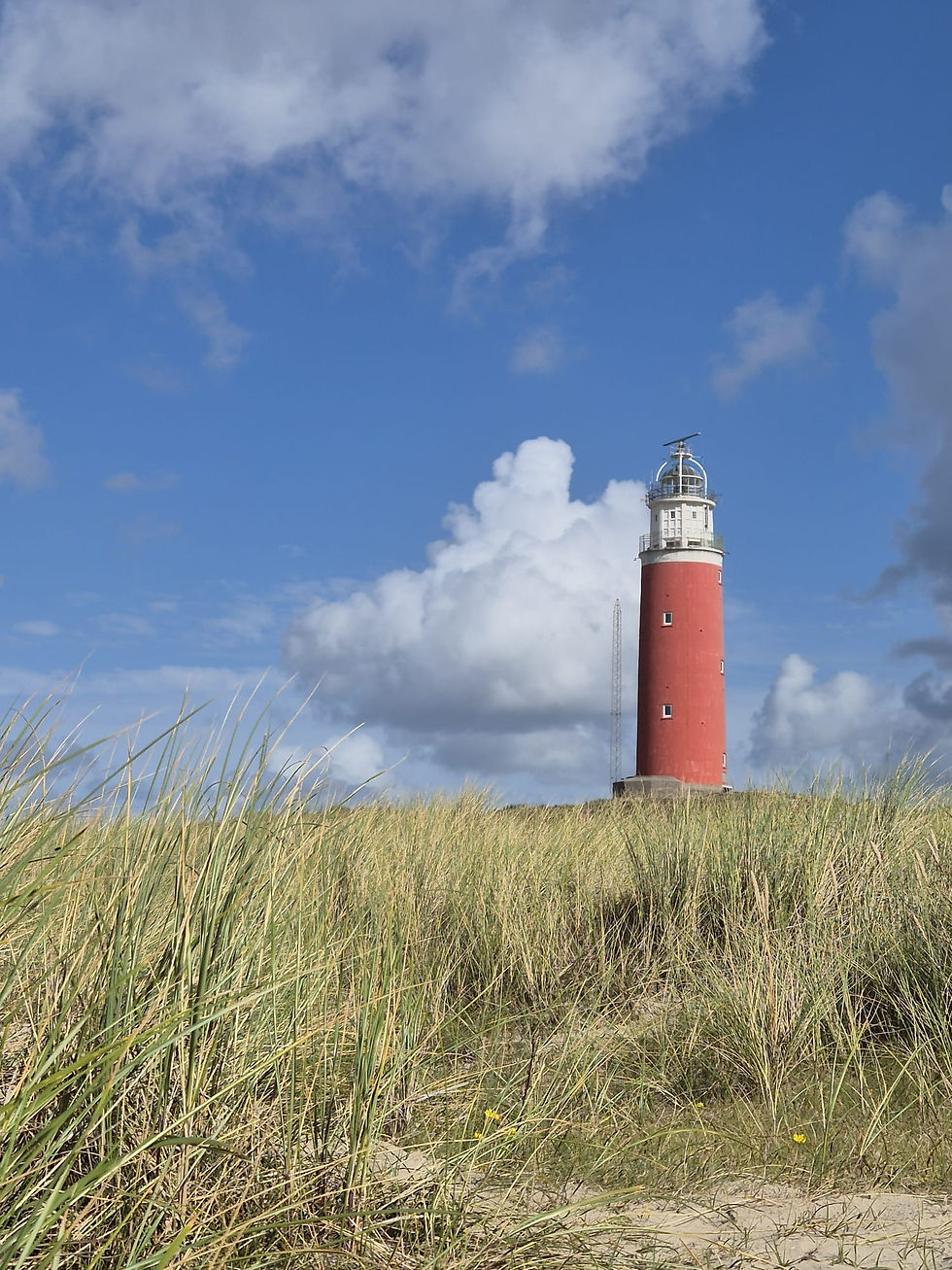 Red lighthouse on sandy dunes under a bright blue sky with large white clouds, calm coastal scene