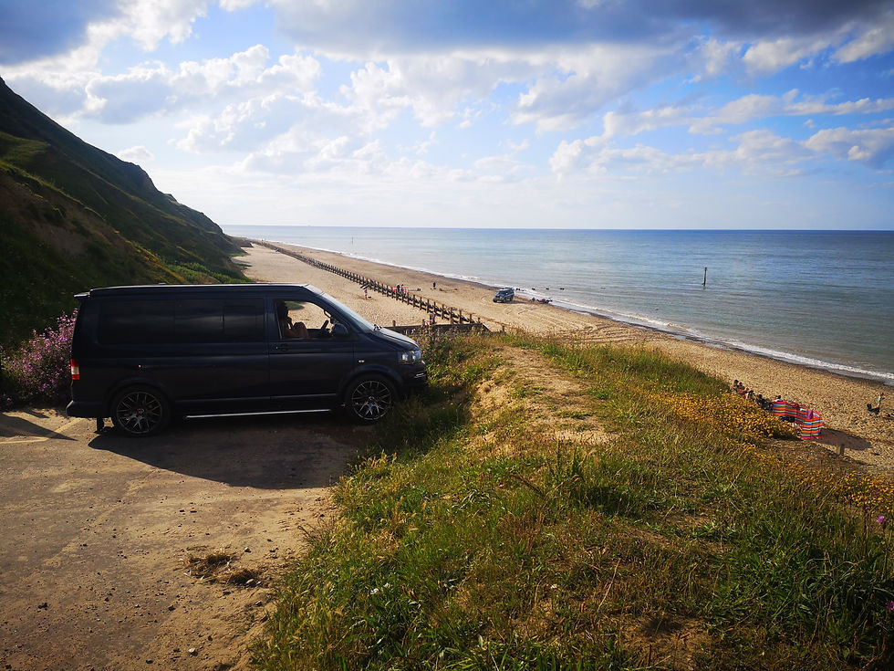 Black van on a cliff above a sandy beach and calm sea under a cloudy blue sky.