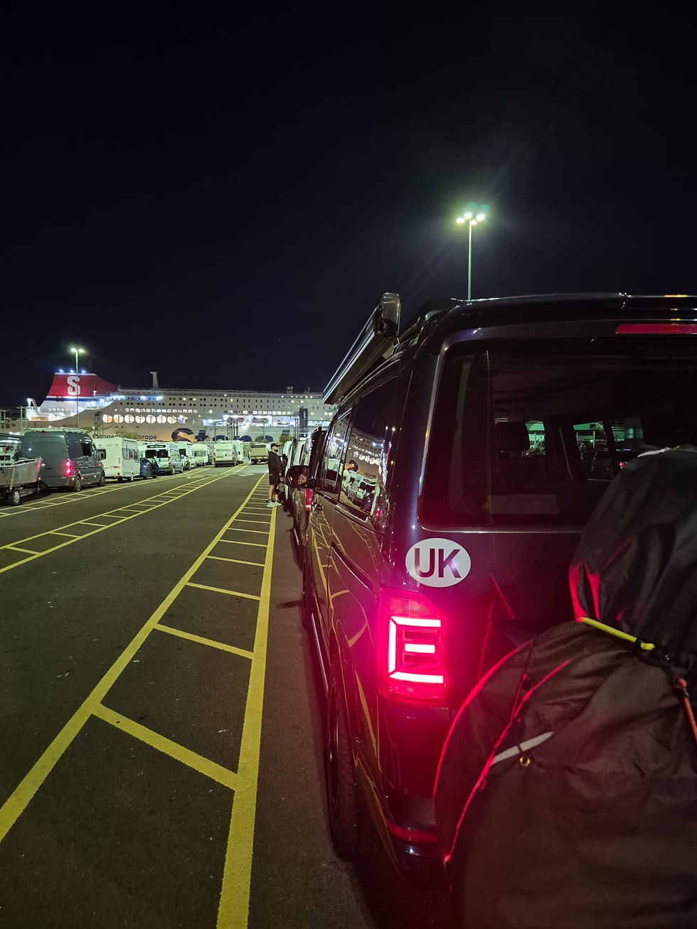 Night ferry terminal with lined-up vans and a large ship; black van with UK sticker and backpack in foreground.