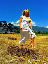 woman with white dress holding a big circle in a field, behind is a mountain, trees, sky